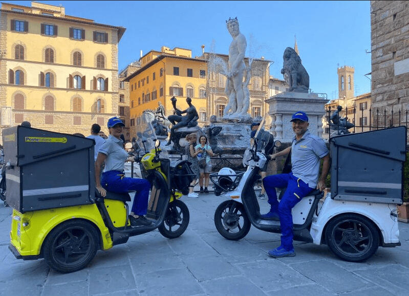 Two Italian mail service employees ride scooters through Florence's historic center, where statues are part of lively urban scenes during an orientation walk.