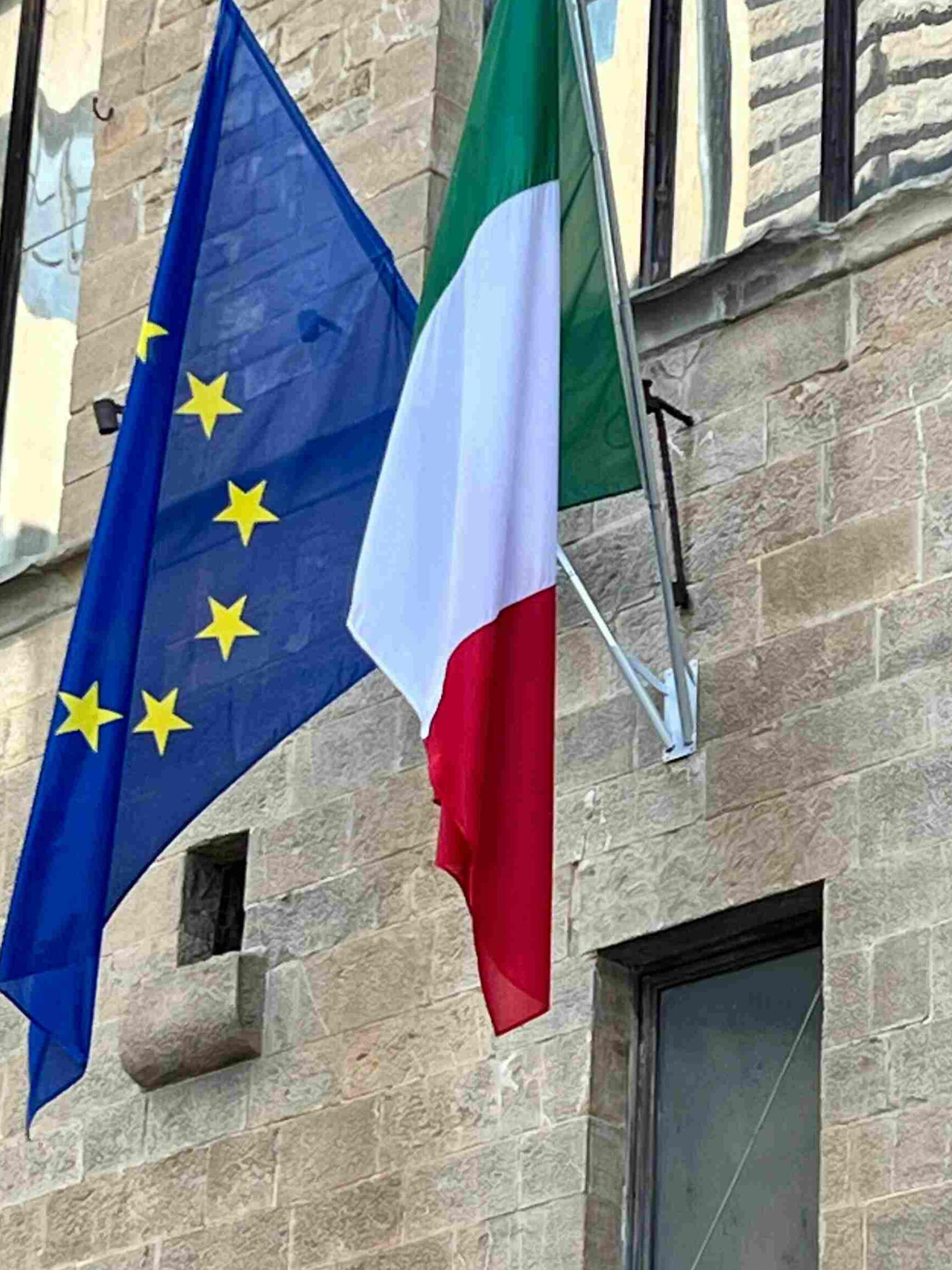 The European Union and Italian flags hang against the stone facade of a historic government building with arched and rectangular windows where Italian visa applications are filed.