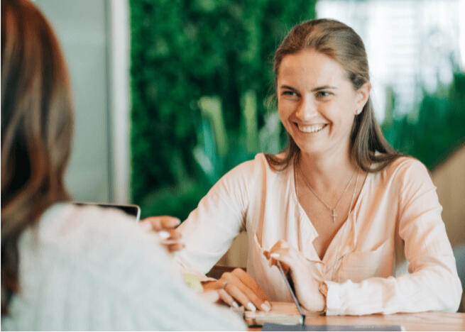 A smiling American woman in a light pink blouse is attending a one-on-one orientation course about daily life in Tuscany. She is sitting across the table from her bilingual coach.
